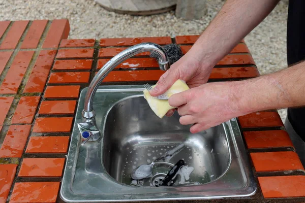 男性の手は石鹸スポンジとシンクの上のフォークを保持します 屋外キッチンでカトラリーを洗う — ストック写真