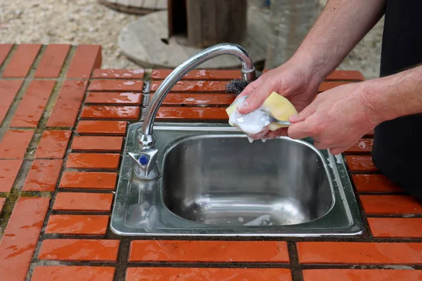 男性の手は シンクの上に石鹸スポンジとスプーンを保持します 屋外キッチンでの食器洗浄 — ストック写真