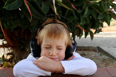 Memnun olmayan altı yaşında, gözleri kapalı bir çocuk. Parkta kulaklıkla müzik dinliyor..