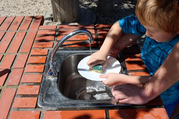子供の手は食器洗いスポンジで皿を洗っている 父親は彼の息子が皿を洗うのを助ける — ストック写真
