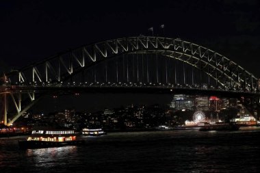 Sydney Liman Köprüsü, Opera Binası 'ndan doğudan batıya doğru seyredilirken ulusal bayraklar ve feribotların dalgalandığı karanlık bir gecede, neredeyse siyah bir gökyüzünün altında ışıldıyor. NSW-Avustralya.