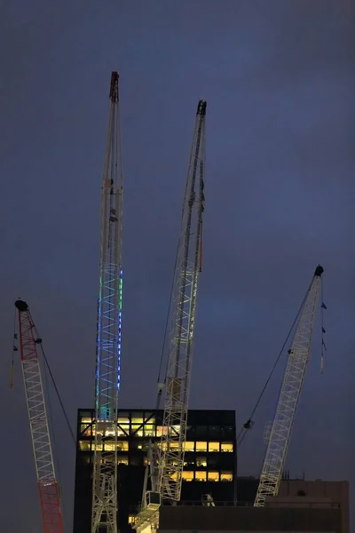 Set of four cranes with pulleys and hanging hooks out of duty placed atop a skyscraper under construction on the CBD viewed at night under dark gray, mostly cloudy sky. Melbourne-VIC-Australia.