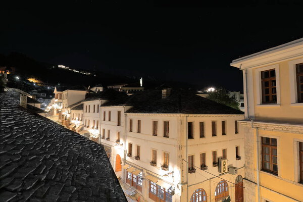 Night view along Rruga Gjin Zenebisi street under a starry sky over an Ottoman-era house roof, covered with flat dressed stones, road climbing up the hill to a telecom tower atop. Gjirokaster-Albania.