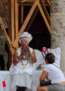 Havana, Küba-7 Ekim 2019: Muhteşem tırnaklı Küba santerası, Plaza Katedral Meydanı 'nın doğu tarafında büyük bir puro içerken kehanet hizmetlerini talep eden bir müşteriye hizmet veriyor..