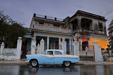Havana, Cuba-October 8, 2019: Blue-white American classic car -almendron- Ford Fairlane 4-door Sedan 1955 with 1956 model's steel side-stripe stationed on Linea Street, wet after heavy afternoon rain. clipart