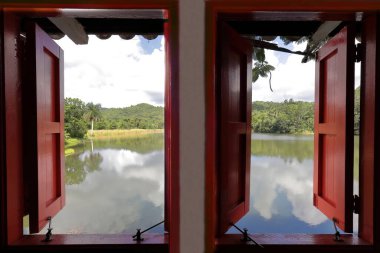 Candelaria, Cuba-October 9, 2019: Red wooden exterior shutters and frame of a double window with open views overlooking Lago San Juan Lake in a local house of Las Terrazas rural tourist eco-community. clipart