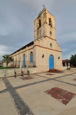 Vinales, Küba-9 Ekim 2019: Krem rengi, mavi kapılı Iglesia del Sagrado Corazon-Sacred Heart Kilisesi - MS 1883 Plaza Central Meydanı 'nda gün batımında gri, bulutlu gökyüzü tehdidi altında.