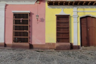 Trinidad, Cuba-October 13, 2019: Refurbished yet already chipped facades of colonial houses on Calle Amargura Street painted yellow and pink featuring brown wood door and grilles with turned balusters clipart