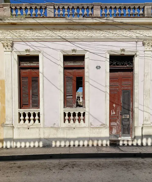 stock image Santiago, Cuba-October 20, 2019: Pale pink wall with white mouldings, chipped maroon-red wood door and window shutters, east-facing facade of uninhabited, dilapidated Eclectic building, Clarin Street.