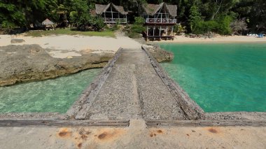 Mayari, Cuba-October 23, 2019: Eastward view from the old pier to the sand strip and the exterior of the thatched, timber-made beach restaurant on Cayo Saetia Cay west shore, Bahia Nipe Bay entrance. clipart