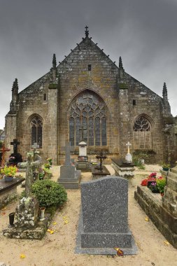 Locronan, France-October 24, 2020: Tombs, graves and tombstones of the local cemetery behind the Saint Ronan Church, its rear facade with the chevet main stained-glass window flanked by two secondary. clipart