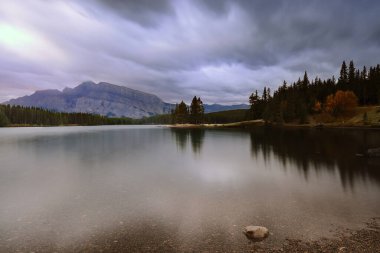 Kuzeybatı kıyısından güneye doğru 2949 metre yüksekliğindeki Rundle Dağı 'na kadar uzanan iki Jack Gölü üzerinde sonbaharın bulutlu şafağında, gökyüzünün renklerini yansıtan yeşil su görülüyor. Banff-Alberta-Kanada.