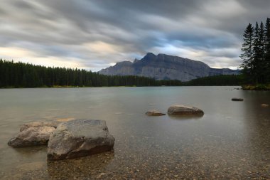 Kuzeybatı kıyısından güneye doğru 2949 metre yüksekliğindeki Rundle Dağı 'na kadar uzanan iki Jack Gölü üzerinde sonbaharın bulutlu şafağında, gökyüzünün renklerini yansıtan yeşil su görülüyor. Banff-Alberta-Kanada.