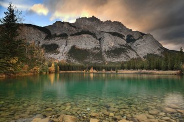 Cascade River ve Pond 'dan 2998 m yüksekliğindeki Şelale Dağı' nın batı-kuzeybatısına doğru sonbahar yapraklarında kozalaklı ağaçlar ve balsam kavak ağaçları üzerinde alacakaranlıkta bulutlu gökyüzü altında. Banff-AB-Kanada.