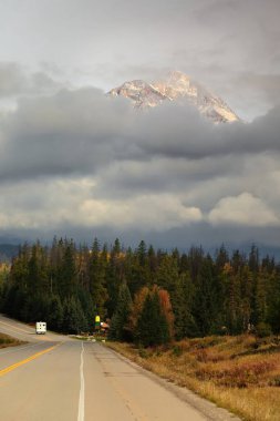 Otoyol AB93-Icefields Parkway, yamaçlarını kaplayan kalın bulutlar ve ayağının altındaki kozalaklı ormanlardan gökyüzüne yükselen güneş ışığıyla aydınlanan Piramit Dağı 'nı işaret ediyor. Jasper NP-Alberta-Kanada.
