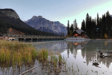 Emerald Gölü 'nün güneybatı ucunda bulutsuz bir şafak ve Emerald Lodge yarımadasına giden yol köprüsü, Emerald, Başkan ve Michael arka planda. Field, Yoho NP, British Columbia, Kanada.