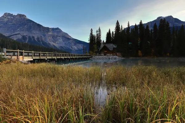 Emerald Gölü 'nün güneybatı ucunda bulutsuz bir şafak ve Emerald Lodge yarımadasına giden yol köprüsü arka planda ise Başkan, Michael ve Wapta. Field, Yoho NP, British Columbia, Kanada.
