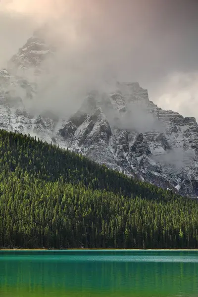 Waputik Sıradağları 'ndaki 3266 metre yüksekliğindeki Chephren Dağı zümrüt yeşili sudan yükselen sis ve Lower Waterfowl Gölü' nün SW kıyısındaki kalın kozalaklı ormandan yükseliyor. Banff NP-Alberta-Kanada.