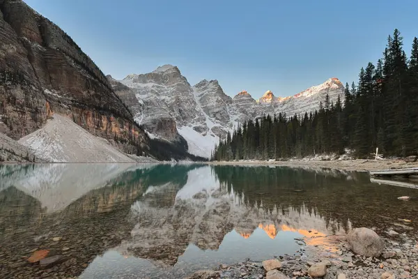 On Tepe Vadisi 'ndeki Moraine Gölü' nün zümrüt yeşili yansıtıcı suları açık mavi gökyüzünün altında bulutsuz bir gündoğumunda taşlı kuzey kıyısından güneybatıya doğru görüldü. Banff NP-Alberta-Kanada.