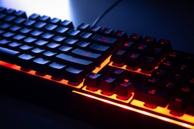 Mekanik RGB klavyesinin yan görüntüsü. Oyun odası modern düzeneği. Kırmızı arka ışık tuşları ve soğuk dramatik ekran ışığı. Programcı ya da bilgisayar oyunu masasının gece görüntüsü. 