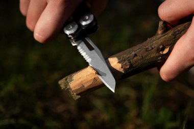 Doğal ışıklandırmada çoklu alet bıçağı kullanarak bir dalı oyan ellerin yakın çekimi. Hayatta kalma, kamp yapma ya da çalılık sahnesi detaylı marangozluk tekniklerini ve doğada maceraya hazır olmayı gösteriyor..