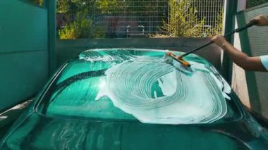 Bir adam oto yıkamada araba yıkar. Genç bir adam arabayı basınç altında köpük ve suyla temizler ve bir bezle siler. Bir arabayı özen ve yaz elle yıkama