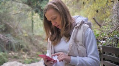 Hamile bir kadın sonbahar parkında oturup akıllı telefondan hamile kadınlara bilgi okuyor. Gelecekteki ebeveynler, anneler ve babalar için çevrimiçi dersler.