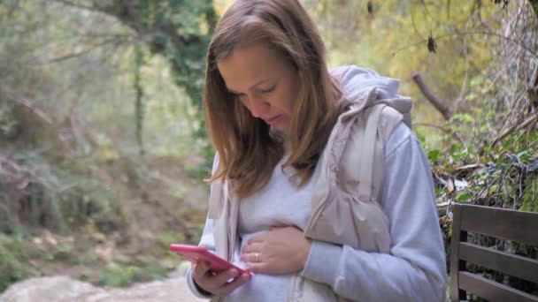 一位孕妇坐在秋天的公园里 用智能手机为孕妇阅读相关信息 为未来的父母 母亲和父亲提供在线课程 — 图库视频影像