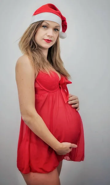 一个孕妇的照片 一个穿着红色衬裙的孕妇 戴着圣诞老人的帽子 双手抱着她的胃 时尚孕妇 — 图库照片