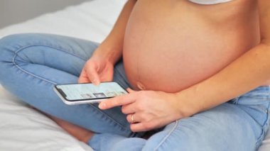 Hamile kadın doğmamış bebek için giysi ve oyuncak seçiyor ve karnını sevgi ve özenle okşuyor. Müstakbel anne online alışveriş için akıllı telefon kullanıyor..