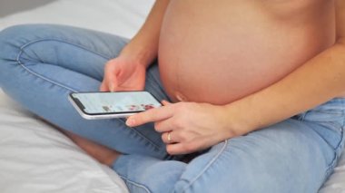 Hamile kadın doğmamış bebek için giysi ve oyuncak seçiyor ve karnını sevgi ve özenle okşuyor. Müstakbel anne online alışveriş için akıllı telefon kullanıyor.