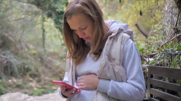 一位孕妇坐在秋天的公园里 用智能手机为孕妇阅读相关信息 为未来的父母 母亲和父亲提供在线课程 — 图库视频影像