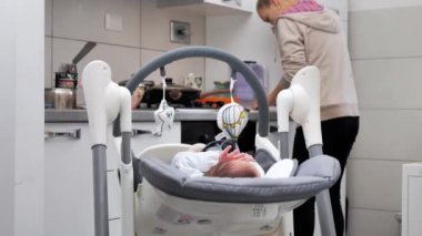 mother is preparing food in the kitchen, the baby is rocking. A housewife performs many tasks at home with a newborn baby.
