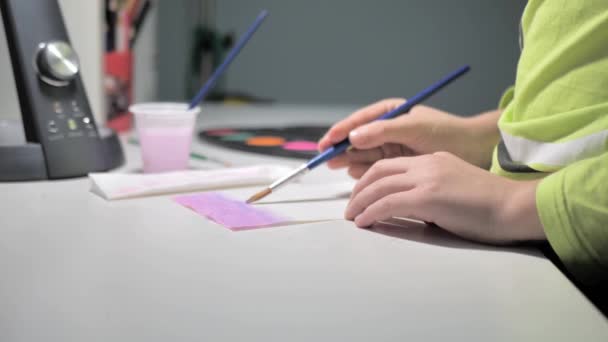 男の子は家の白いテーブルに座っている間にブラシとピンクの塗料でペイントします 少年は水彩画で絵を描くことを学ぶ 少年が美術の第一歩を踏み出す — ストック動画