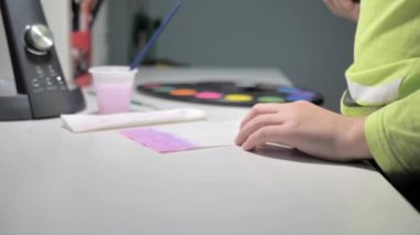 boy paints with a brush and pink paint while sitting at a white table at home. The boy learns to paint with watercolors. A boys first steps in fine art.