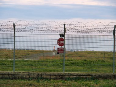 Havaalanı güvenlik çit sistemi, dikenli telle çevrili, güvenlik yasaklı bölge dur levhası ve arkada uçaklar var..