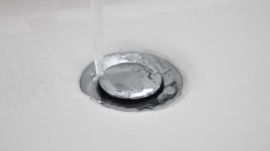 Su lavabo deliğinden aşağı akıyor. Seramik lavaboda yavaş çekim su gideri. Su akıntısı kanalizasyona akıyor, kapatın. Su akmaya başlar ve görüntülerin sonunda biter. Kamu hizmetleri. kanalizasyon.