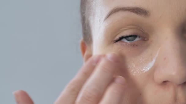 若い女性のクローズアップビデオ撮影は彼女のチェックに保湿剤を持っています 彼女は彼女の指のスキンケアの概念で彼女の顔に製品をマッサージ健康肌の治療 — ストック動画