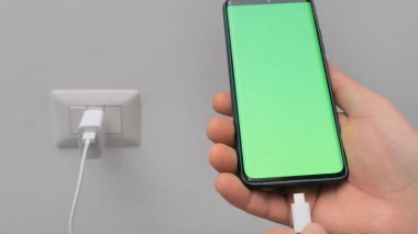Erkek eliyle şarj cihazına bağlı yeşil ekranlı bir telefon. Bir el akıllı telefonu tutar ve onu şarj etmeye hazırlanır..