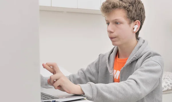 子供は机の上でノートパソコンを使う 巻き毛の少年はヘッドフォンを着用している音楽を聞いて コンピュータゲームをして インターネットを閲覧して 友人とチャットしている — ストック写真
