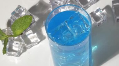 Bir adamın eli bir kokteylin içine pipet koyar, beyaz arka planda buzlu ve naneli mavi bir kokteyl.