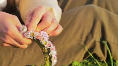 Bir kadın çimenlerde oturur ve papatya çelengi örer..