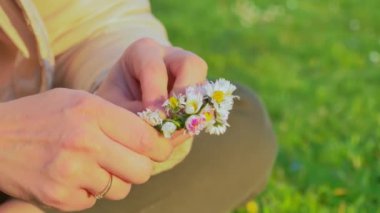 Güneş ışığıyla yıkanmış canlı bir çayırda bir çift el, zarif çiçekleri ustalıkla büyüleyici bir taç gibi dokuyor. Yumuşak yapraklar bir renk paleti sergilerken, yeşillik doğal bir dokunuş ekliyor.