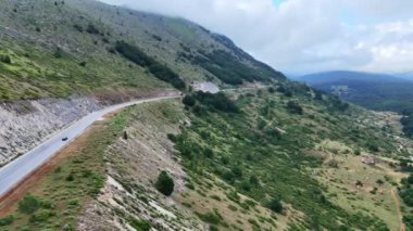 Büyüleyici bir manzara, dağlık arazinin çevresini zarifçe saran dolambaçlı bir yol ile gözler önüne seriliyor. Canlı yeşillikler tepeleri örtüyor, bulutlu gökyüzüne karşı çarpıcı bir kontrastlık yaratıyor.