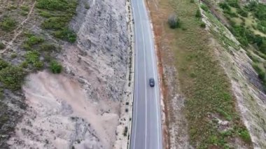 Yalnız bir araç engebeli bir dağ yamacının çevresini saran dolambaçlı bir yoldan geçiyor. Manzara, kayalık arazi ve canlı yeşilliğin bir karışımını gösteriyor. Günden akşama geçişi simgeliyor.
