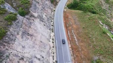Şık bir araba, yemyeşil ve kayalık tepelerle çevrili, dolambaçlı bir dağ yolunda zarifçe süzülüyor. Gökyüzü yumuşak bulutlarla süslenir ve manzaraya yumuşak gölgeler bırakır. Doğaldır