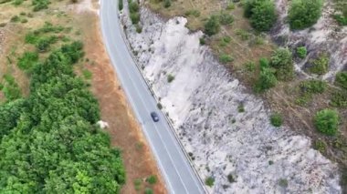 Bir araba, dalgalı yeşilliklerle çevrili kayalık bir yamacın kenarını saran dolambaçlı bir yolda yol alır. Güneş ışığı, engebeli arazi ile bereketli arazi arasındaki zıtlığı vurguluyor.