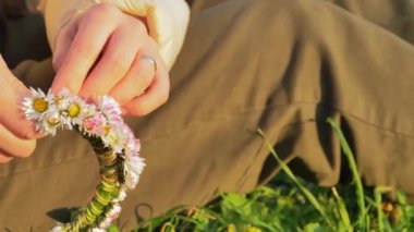 Bir insan yemyeşil bir çayırda oturur ve çarpıcı bir tacı narin çiçeklerle örer. Altın güneş ışığı yaprakların arasından süzülerek canlı çiçeklerin üzerine sıcak bir parıltı saçar. Atmosfer sakin.