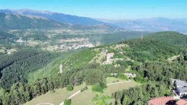 Yükselmiş bir bakış açısından bakıldığında, çarpıcı bir manzara gözler önüne seriliyor, ağaçlarla örtülü yeşil tepeler, aralarında büyüleyici bir köy bulunuyor. Açık mavi gökyüzü dinginliği tamamlıyor