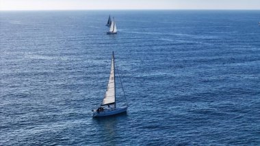 Üç zarif yelkenli sakin sularda yüzüyor, beyaz yelkenleri derin mavi denize karşı güzel bir tezat oluşturuyor. Öğleden sonra güneşi sahnenin huzurunu vurguluyor..